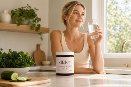 Woman holding a glass of water at a sunlit kitchen counter with fresh greens and cucumber nearby, morning wellness routine for akkermansia and blood sugar health