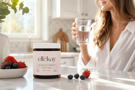 Woman in a bright morning kitchen holding a glass of water with fresh fruit on a white marble counter, representing digestive wellness